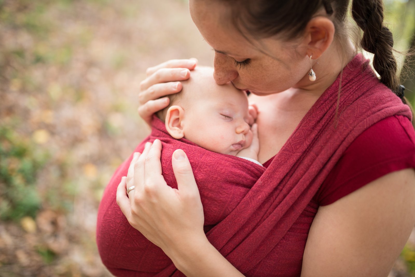 Matka nosiaca svoje dieťa v šatke, bozkávajúca na čelo vonku počas jesene